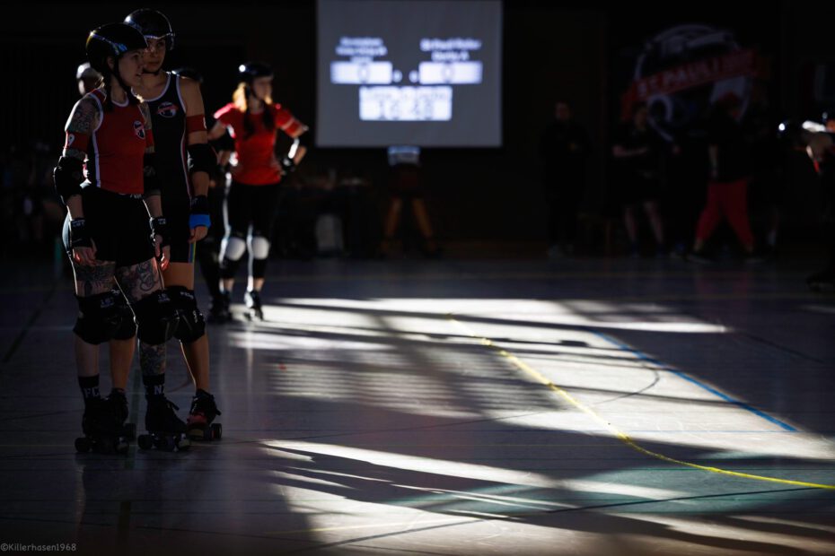 Auf dem Track stehen im Schatten mehrere Roller Derby Spieler:innen. Im Hintergrund steht das Scoreboard.